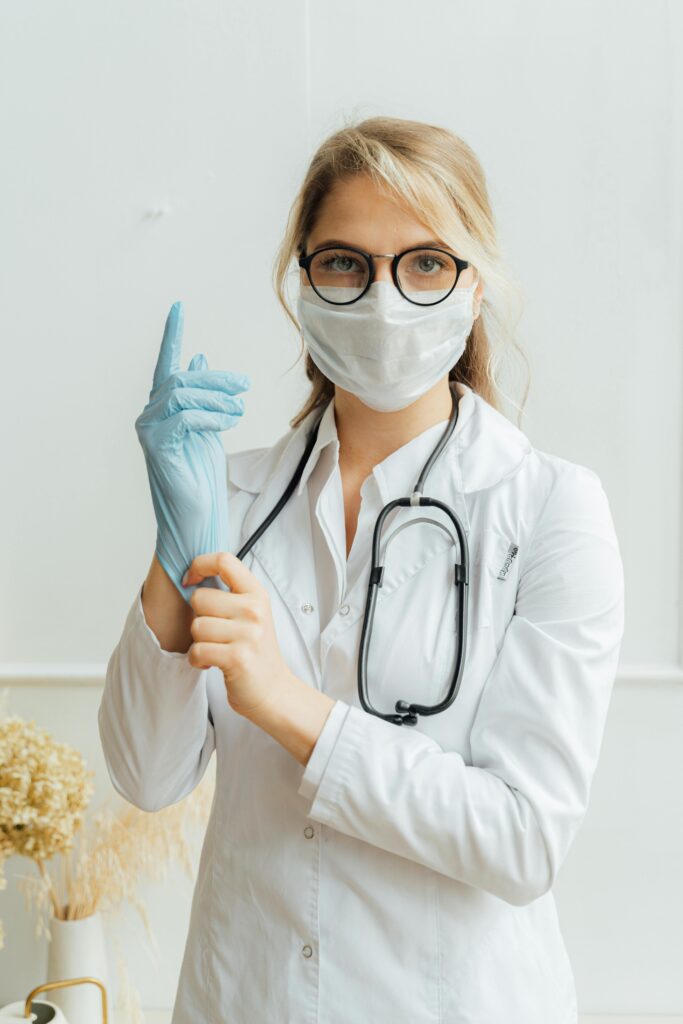 pexels photo 5214949 5214949 Female doctor wearing eyeglasses, face mask, and stethoscope, putting on latex gloves indoors.